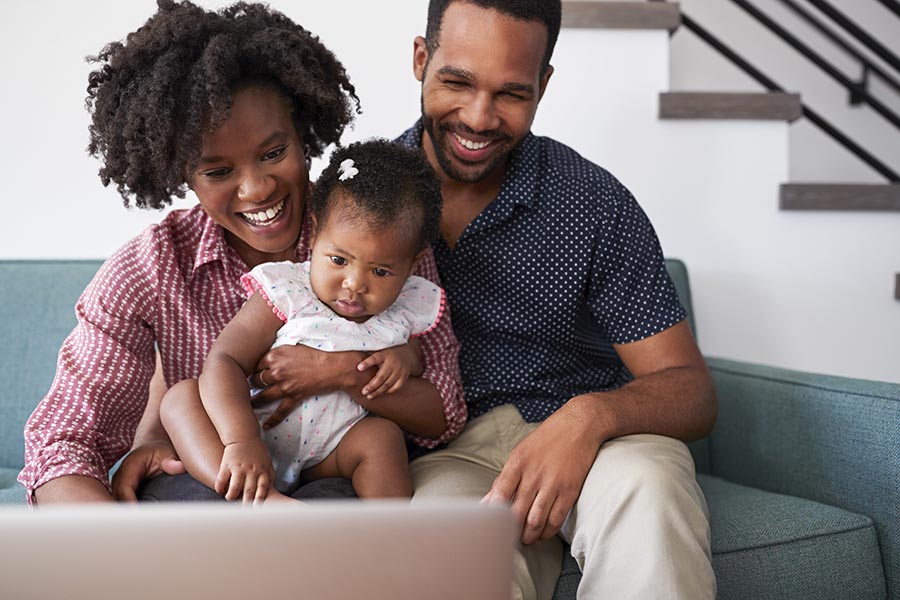 Client Center - a Mother and Father and Baby Sitting on Their Light Blue Couch, Using Their Computer, All Happy and Smiling